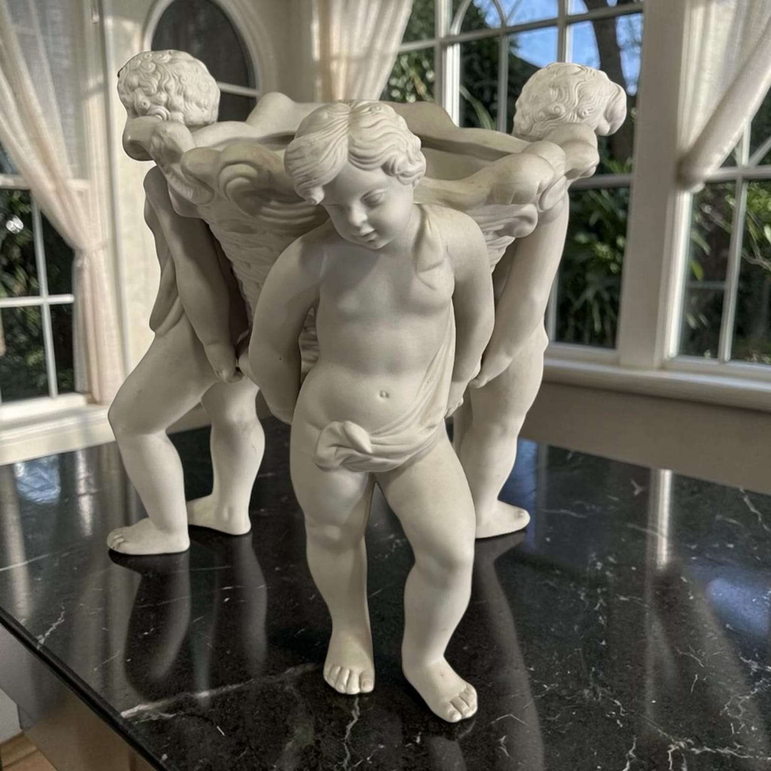 Escultura clásica de porcelana con querubines y figura femenina, decorativa y elegante para mesas y vitrinas, en tonos blancos.