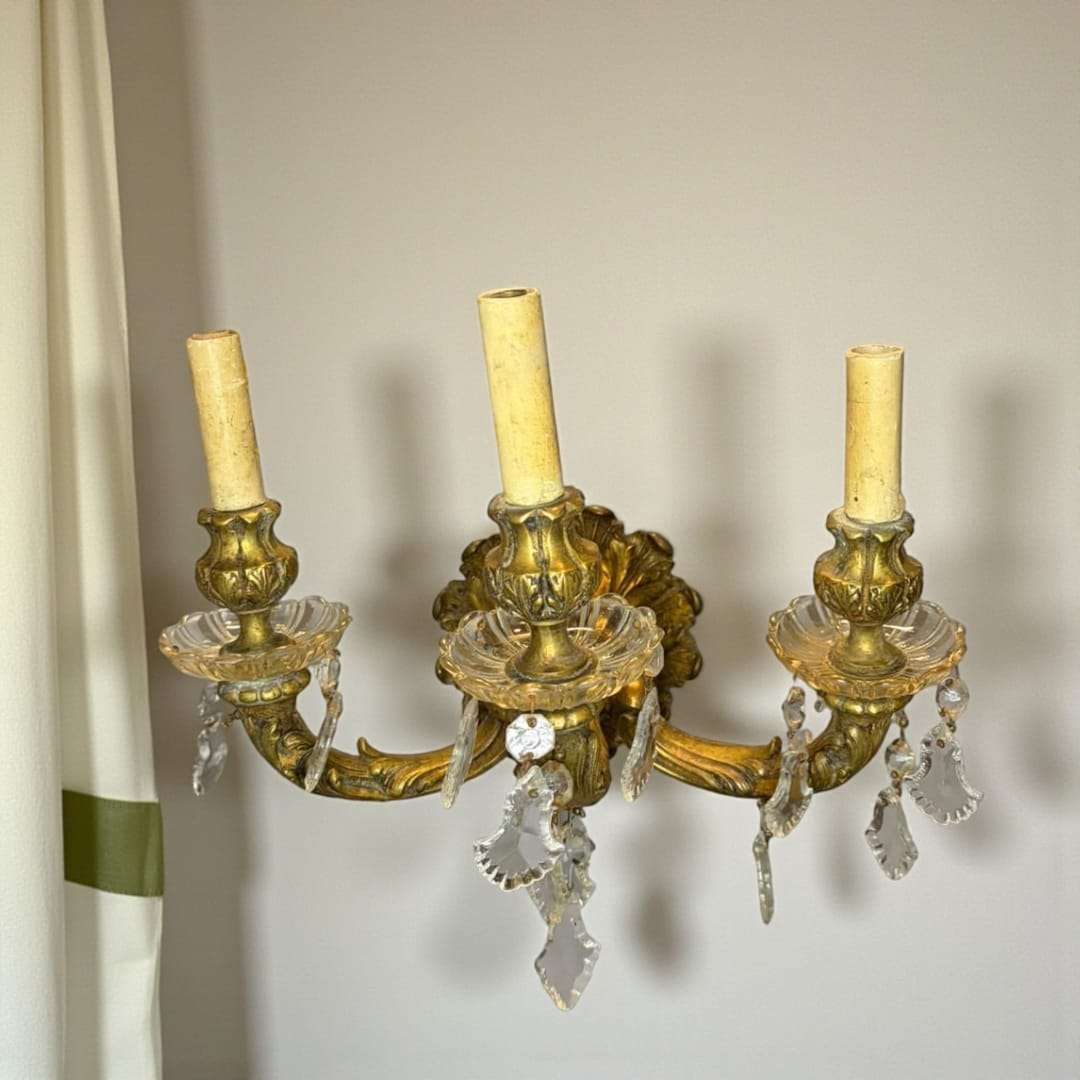 Su estructura, de 32 cm de ancho, presenta tres brazos ornamentales en bronce envejecido, cuidadosamente moldeados con motivos vegetales y curvas arm