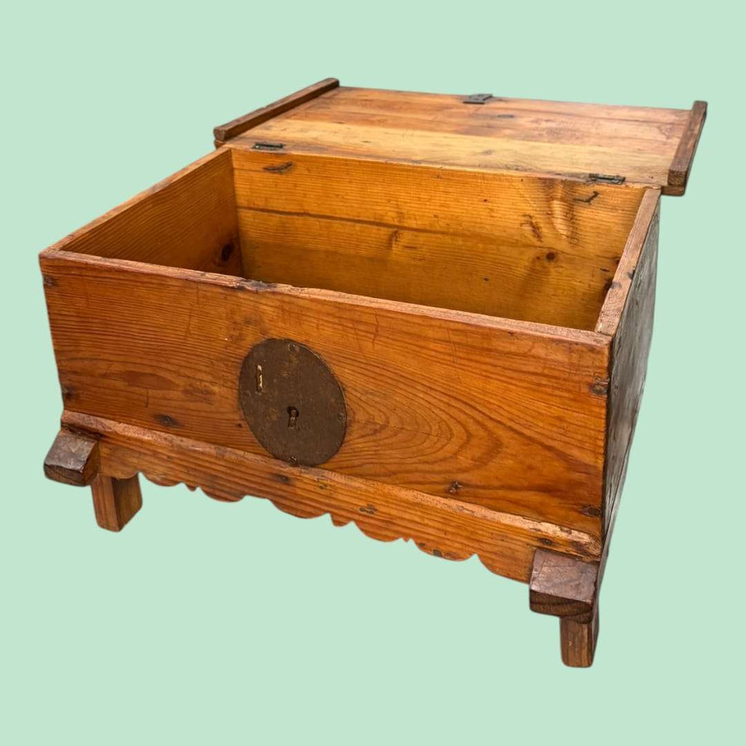 
El cofre de madera presenta una estructura robusta, con paredes gruesas y una tapa superior abatible mediante bisagras metálicas originales, lo que garantiza estabilidad y durabilidad.