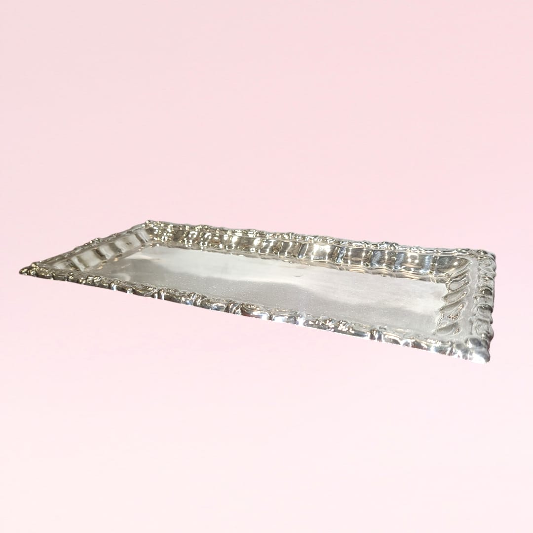Bandeja rectangular de metal plateado con bordes decorativos, rodeada de rosas rojas sobre fondo claro. Ideal para decoración elegante.