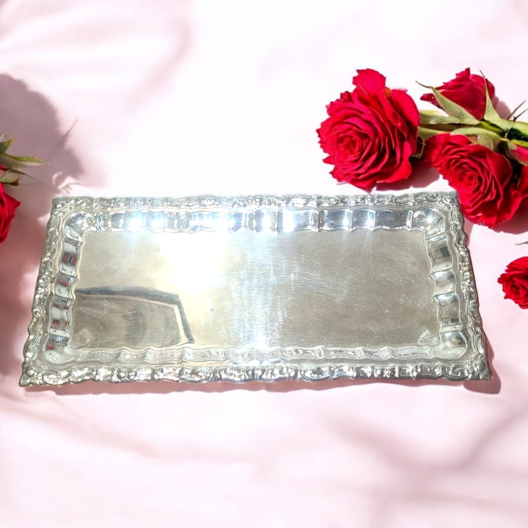 Bandeja rectangular de metal plateado con bordes decorativos, rodeada de rosas rojas sobre fondo claro. Ideal para decoración elegante.