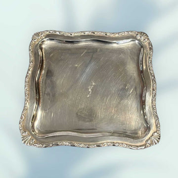 Bandeja de metal plateado con patas y bordes decorativos en forma cuadrada, ideal para decoración vintage en el hogar o jardín.