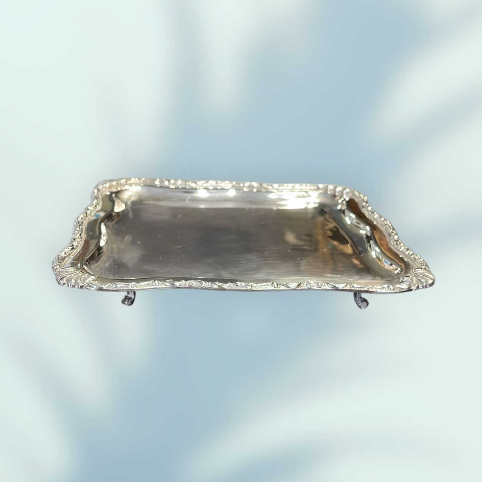 Bandeja de metal plateado elevada con patas y bordes ornamentados, perfecta para servir o como pieza decorativa exclusiva.