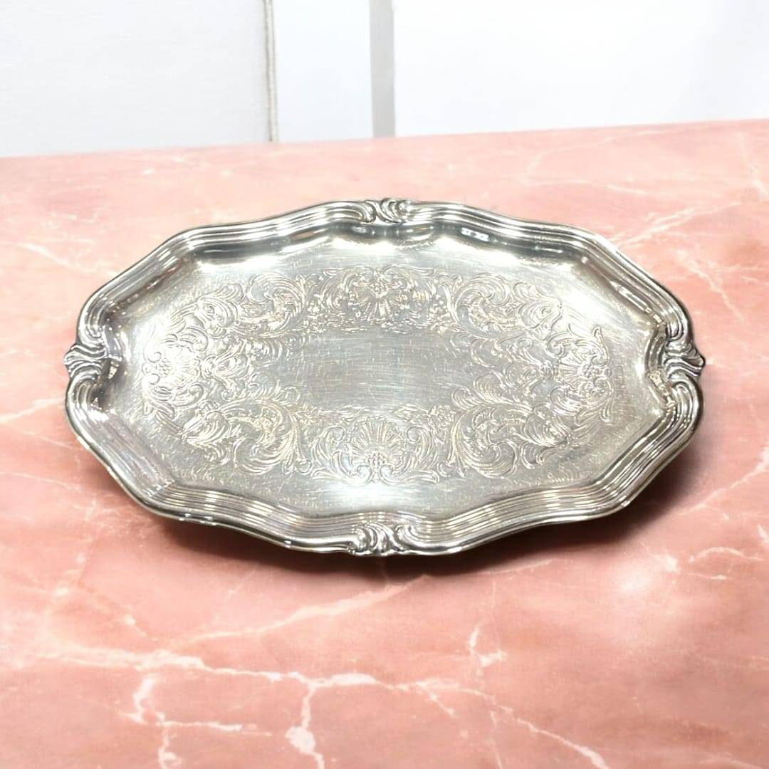 Bandeja elegante de metal plateado con grabados vintage ornamentales sobre superficie rosada. Ideal para decoración clásica y coleccionismo.
