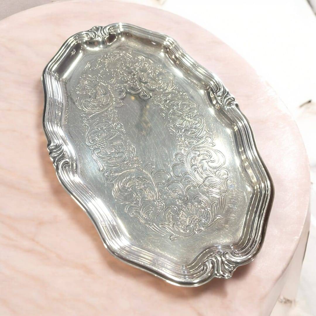 Bandeja elegante de metal plateado con grabados vintage ornamentales sobre superficie rosada. Ideal para decoración clásica y coleccionismo.