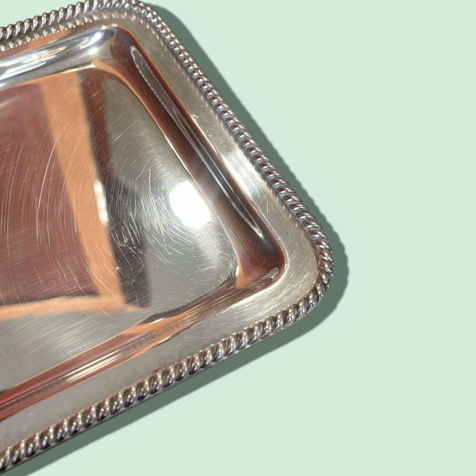 Detalle del borde trenzado repujado de la bandeja de metal plateado, estilo clásico perfecto para decoración sofisticada.