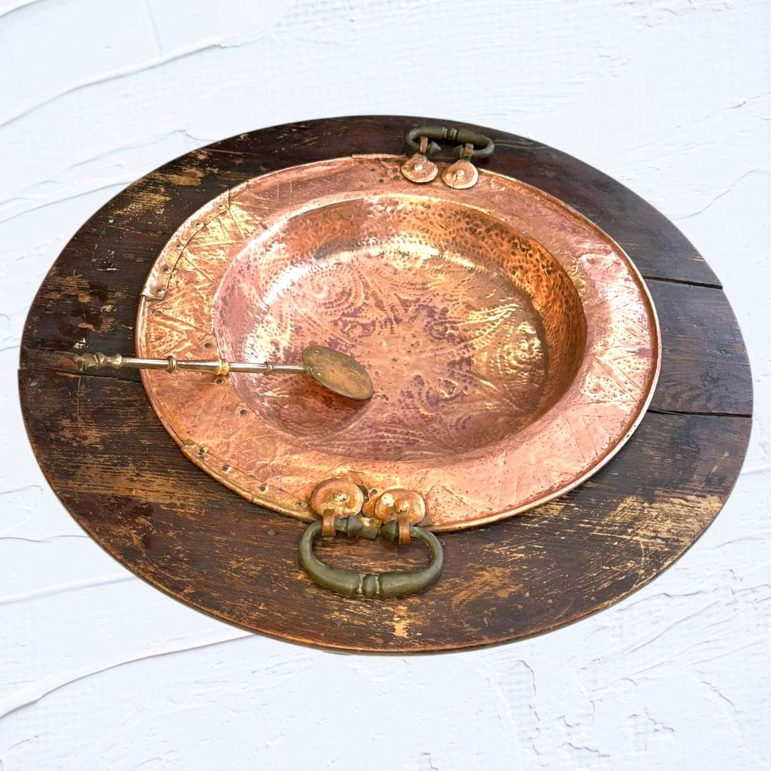 Brasero de cobre martillado con remaches visibles, base circular de madera oscura y badila antigua. Perfecto para decoración vintage en interiores.
