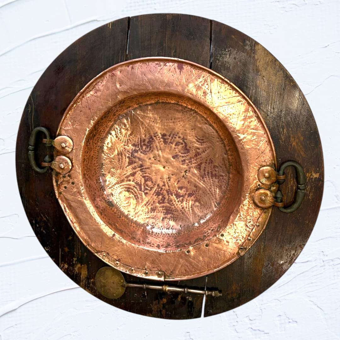 Brasero de cobre con detalles grabados, montado sobre base de madera circular y acompañado de badila. Pieza decorativa única para el hogar.