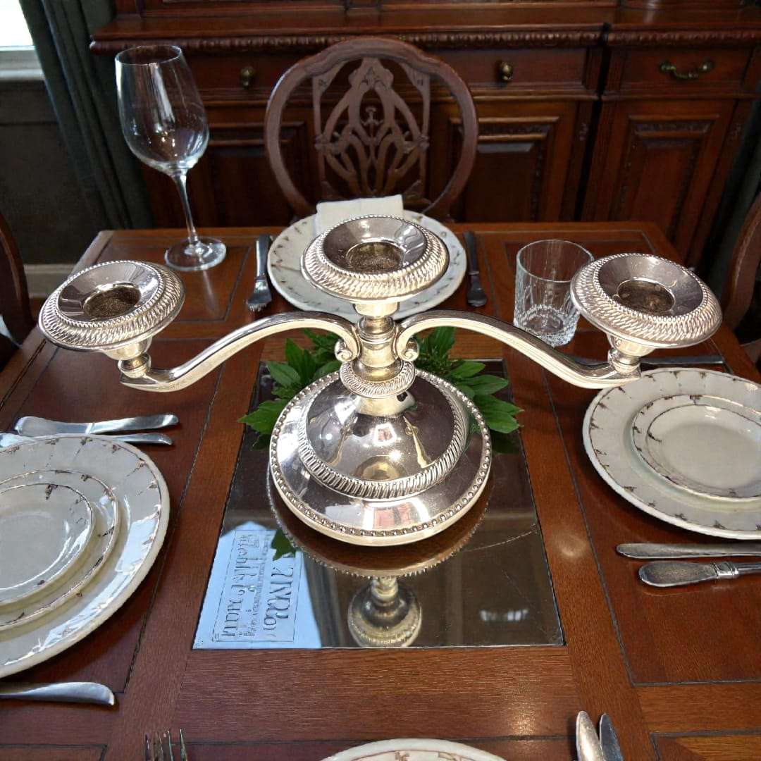 Candelabro de tres velas en metal plateado vintage sobre mesa elegante con vajilla clásica. Ideal para decoración de cenas y eventos formales.