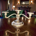 Candelabro de tres velas en metal plateado vintage con diseño clásico sobre mesa de madera. Ideal para decoración de interiores elegantes.