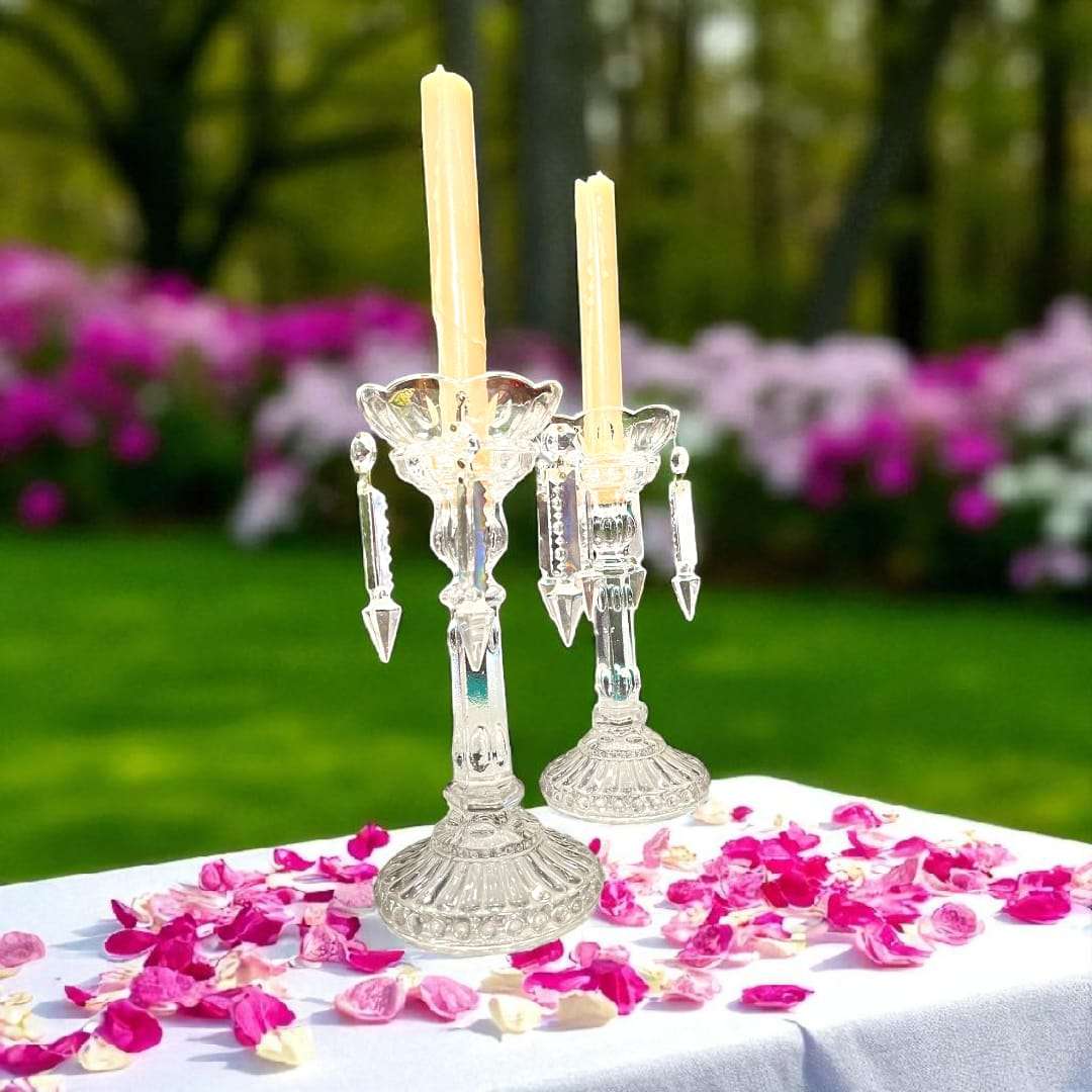 Pareja de candeleros de cristal antiguos sobre mesa con pétalos, ideales para decoración elegante y tradicional en jardín primaveral.