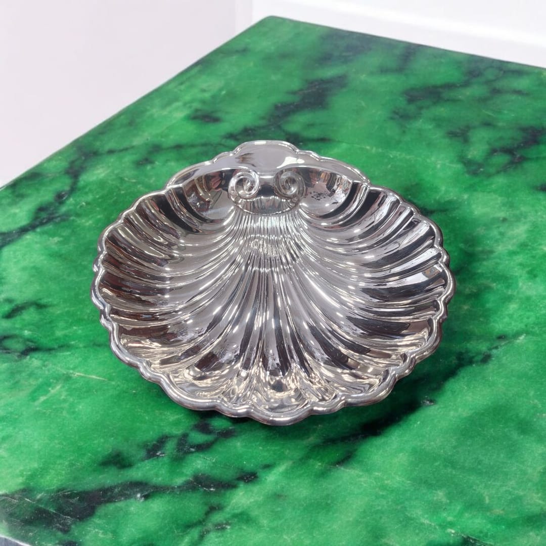 Centro de mesa de metal plateado con forma de concha marina sobre superficie de mármol verde, resaltando su brillo vintage.