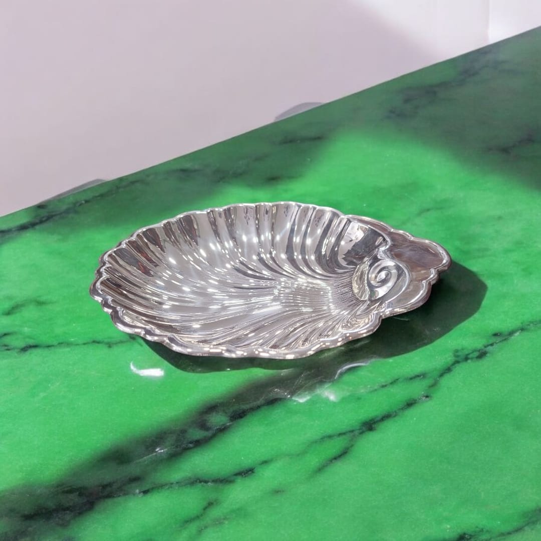 Centro de mesa de metal plateado con forma de concha marina sobre superficie de mármol verde, resaltando su brillo vintage.