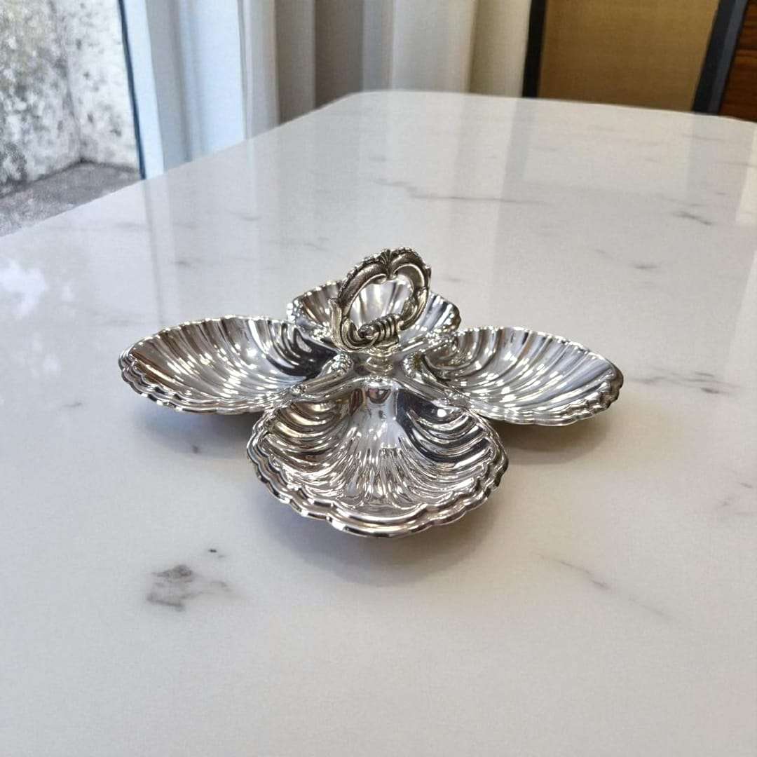 Entremecero antiguo de metal plateado con diseño de conchas y asa central, pieza decorativa funcional para servir en reuniones elegantes.