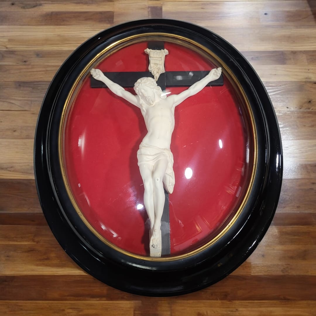 Vista frontal del crucifijo antiguo con cúpula de cristal y marco ovalado en negro. La figura de Cristo tallada en barro de pipa resalta sobre el fondo de terciopelo rojo, creando un efecto visual impactante.