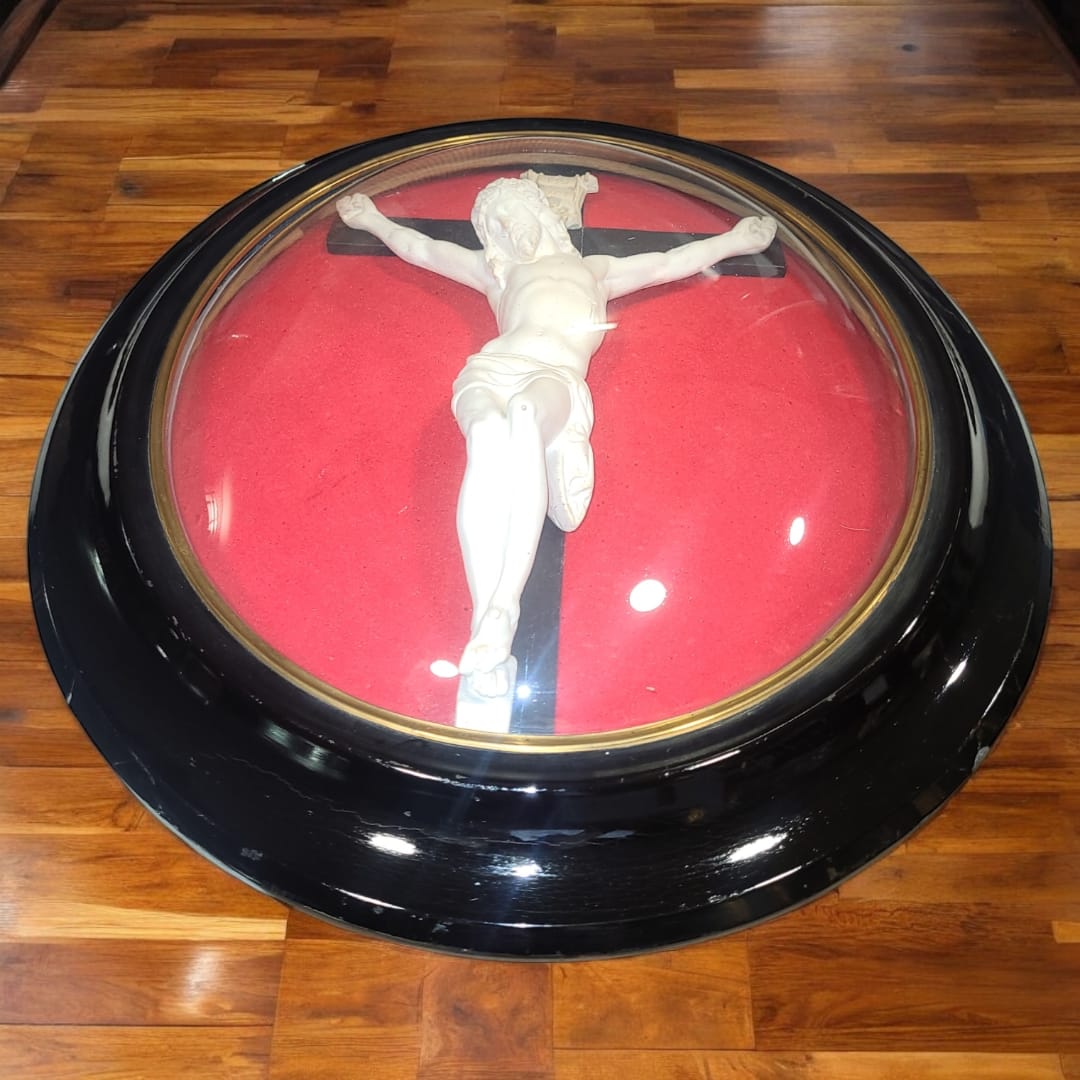 Vista frontal del crucifijo antiguo con cúpula de cristal y marco ovalado en negro. La figura de Cristo tallada en barro de pipa resalta sobre el fondo de terciopelo rojo, creando un efecto visual impactante.