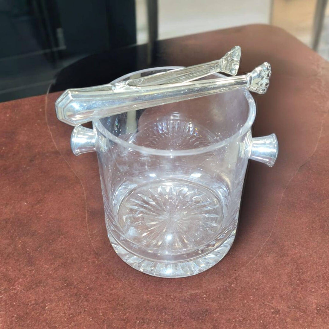 Cubitera de cristal tallado con asas laterales y pinzas de metal plateado sobre mesa marrón. Ideal para decorar y servir hielo con estilo.