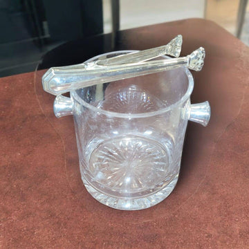 Cubitera de cristal tallado con asas laterales y pinzas de metal plateado sobre mesa marrón. Ideal para decorar y servir hielo con estilo.