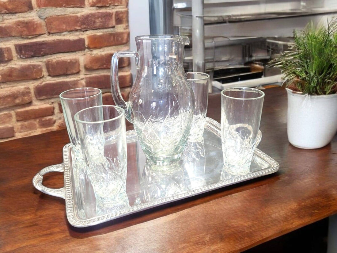 Juego de jarra de cristal y vasos tallados, elegante y clásico, sobre bandeja metálica decorativa en mesa de madera rústica. Ideal para decoración de cocina o comedor.