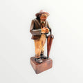 Figura de hombre mayor en madera con sombrero y paraguas, estilo rústico europeo. Pieza decorativa artesanal para espacios clásicos.