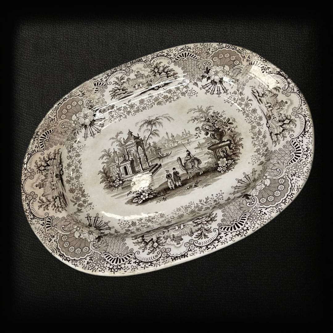 La fuente de loza La Cartuja Pickman es una joya de la cerámica andaluza del siglo XIX, fabricada en la prestigiosa manufactura sevillana La Cartuja de Sevilla, fundada por Charles Pickman. Esta pieza excepcional destaca por su decoración transferware en tono negro sepia, que representa un paisaje orientalista con figuras, arquitectura clásica y ornamentación floral, todo enmarcado por un delicado borde decorado con motivos vegetales.

Con su estilo refinado y su impecable conservación, esta fuente de loza 