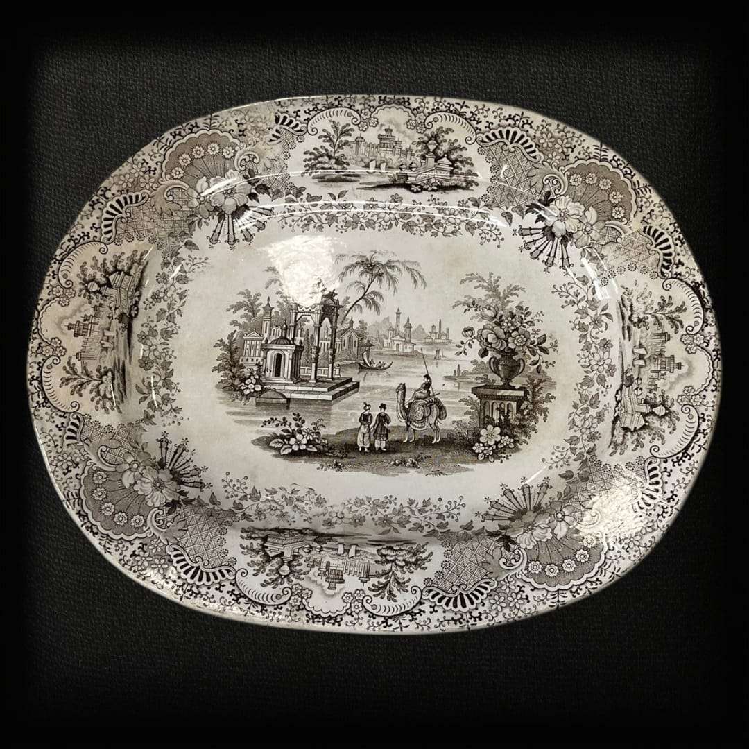 La fuente de loza La Cartuja Pickman es una joya de la cerámica andaluza del siglo XIX, fabricada en la prestigiosa manufactura sevillana La Cartuja de Sevilla, fundada por Charles Pickman. Esta pieza excepcional destaca por su decoración transferware en tono negro sepia, que representa un paisaje orientalista con figuras, arquitectura clásica y ornamentación floral, todo enmarcado por un delicado borde decorado con motivos vegetales.

Con su estilo refinado y su impecable conservación, esta fuente de loza 