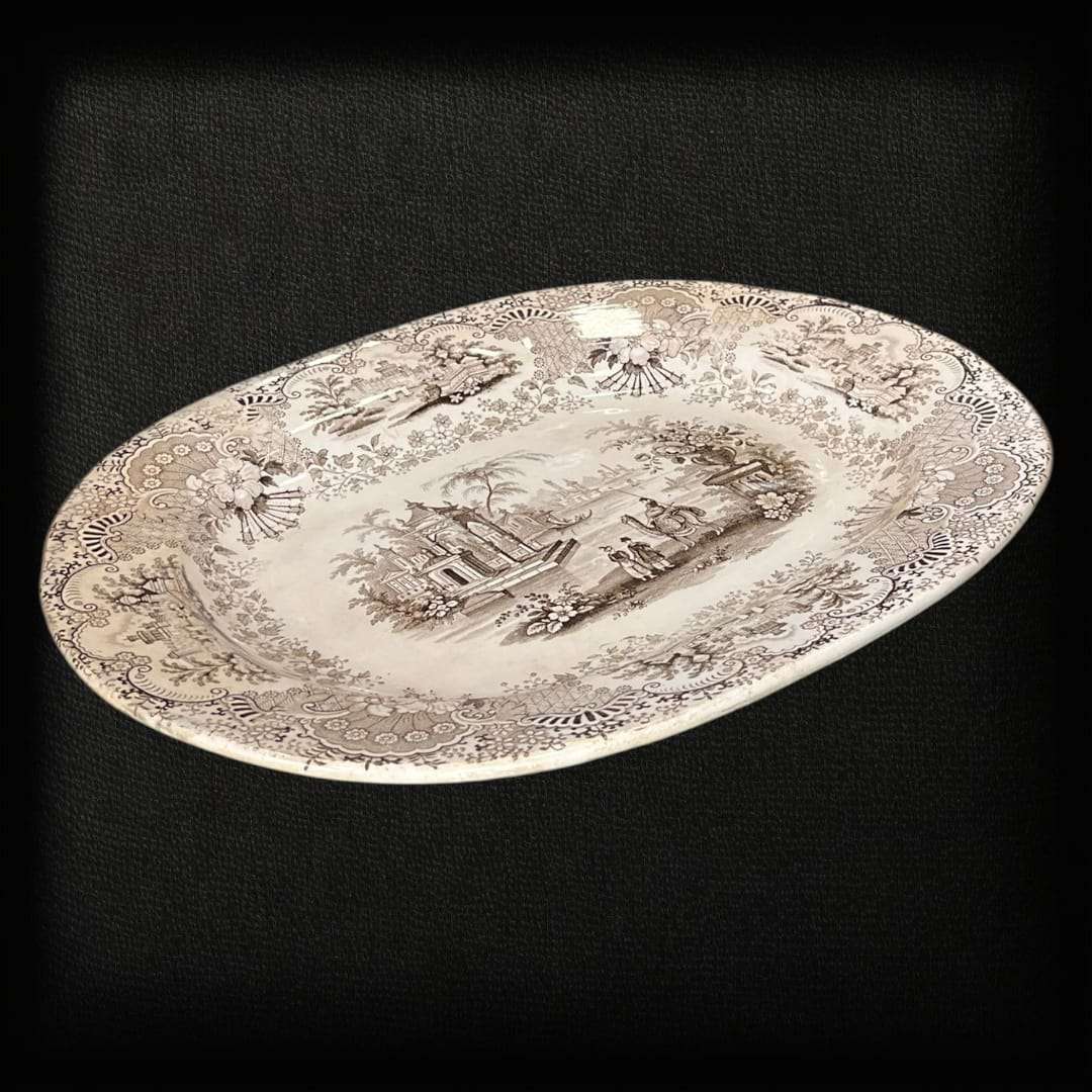 La fuente de loza La Cartuja Pickman es una joya de la cerámica andaluza del siglo XIX, fabricada en la prestigiosa manufactura sevillana La Cartuja de Sevilla, fundada por Charles Pickman. Esta pieza excepcional destaca por su decoración transferware en tono negro sepia, que representa un paisaje orientalista con figuras, arquitectura clásica y ornamentación floral, todo enmarcado por un delicado borde decorado con motivos vegetales.

Con su estilo refinado y su impecable conservación, esta fuente de loza 