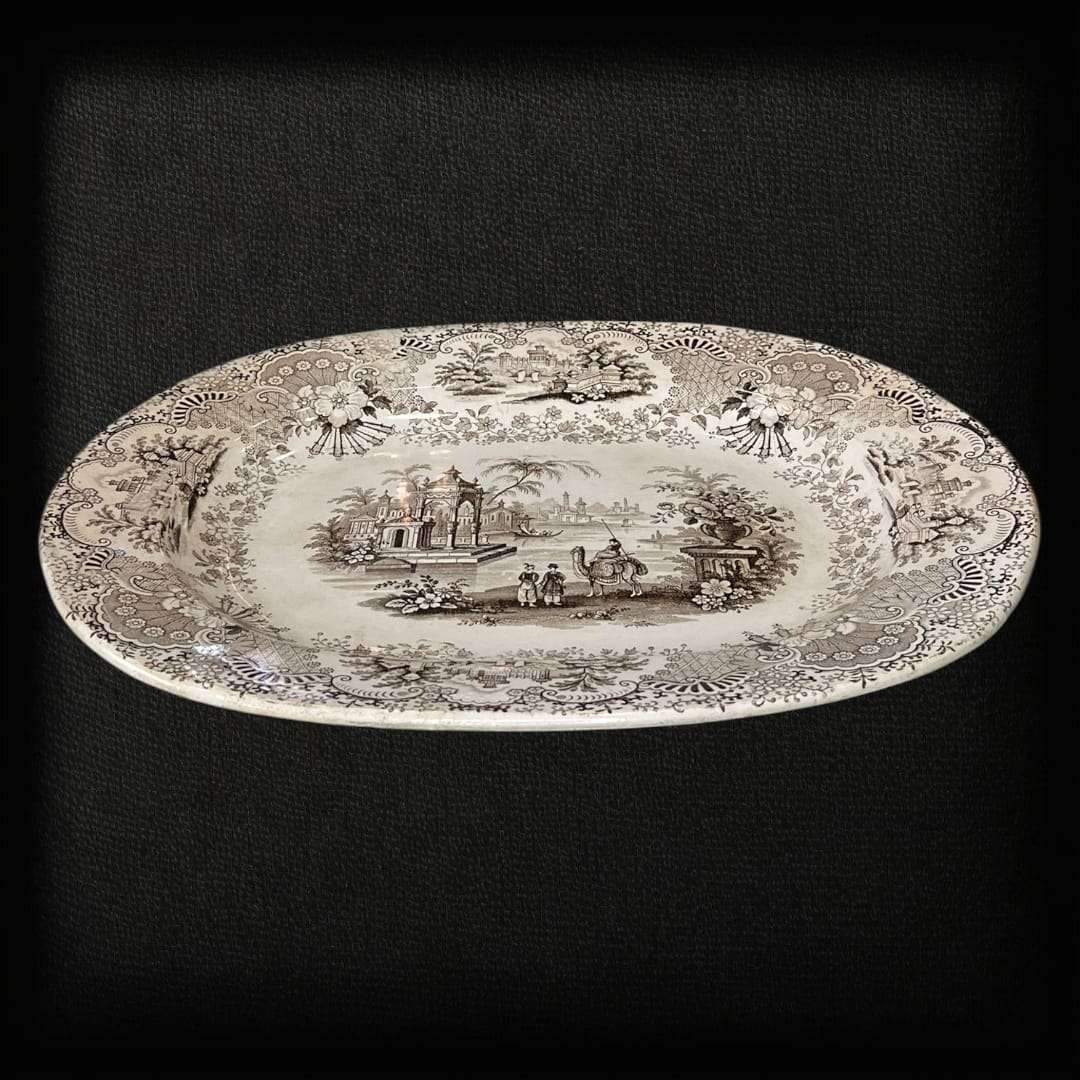 La fuente de loza La Cartuja Pickman es una joya de la cerámica andaluza del siglo XIX, fabricada en la prestigiosa manufactura sevillana La Cartuja de Sevilla, fundada por Charles Pickman. Esta pieza excepcional destaca por su decoración transferware en tono negro sepia, que representa un paisaje orientalista con figuras, arquitectura clásica y ornamentación floral, todo enmarcado por un delicado borde decorado con motivos vegetales.

Con su estilo refinado y su impecable conservación, esta fuente de loza 