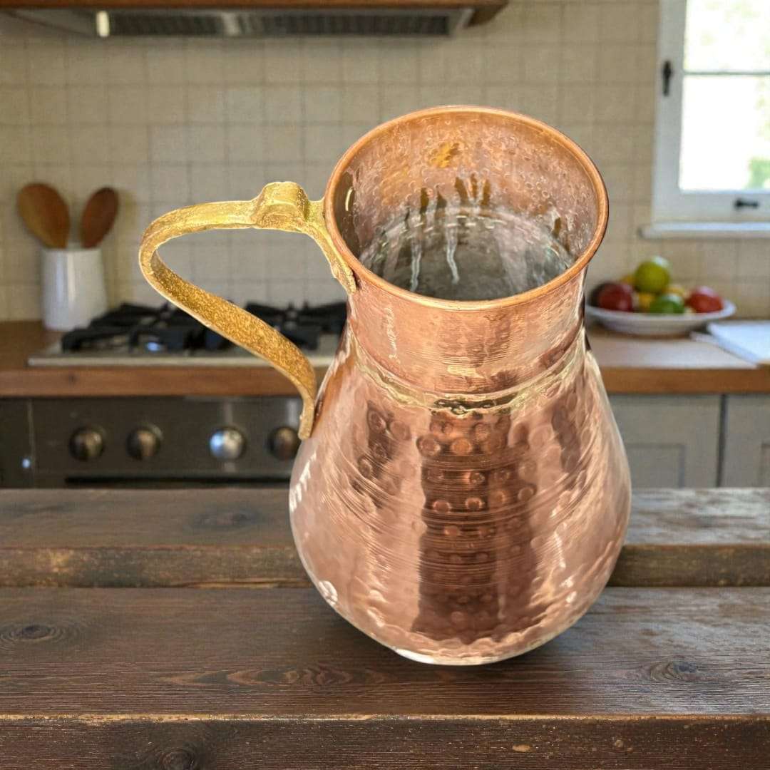 
La jarra de cobre con asa de metal destaca por su cuerpo abombado y cuello ligeramente estrechado, diseño pensado originalmente para conservar líquidos como agua
