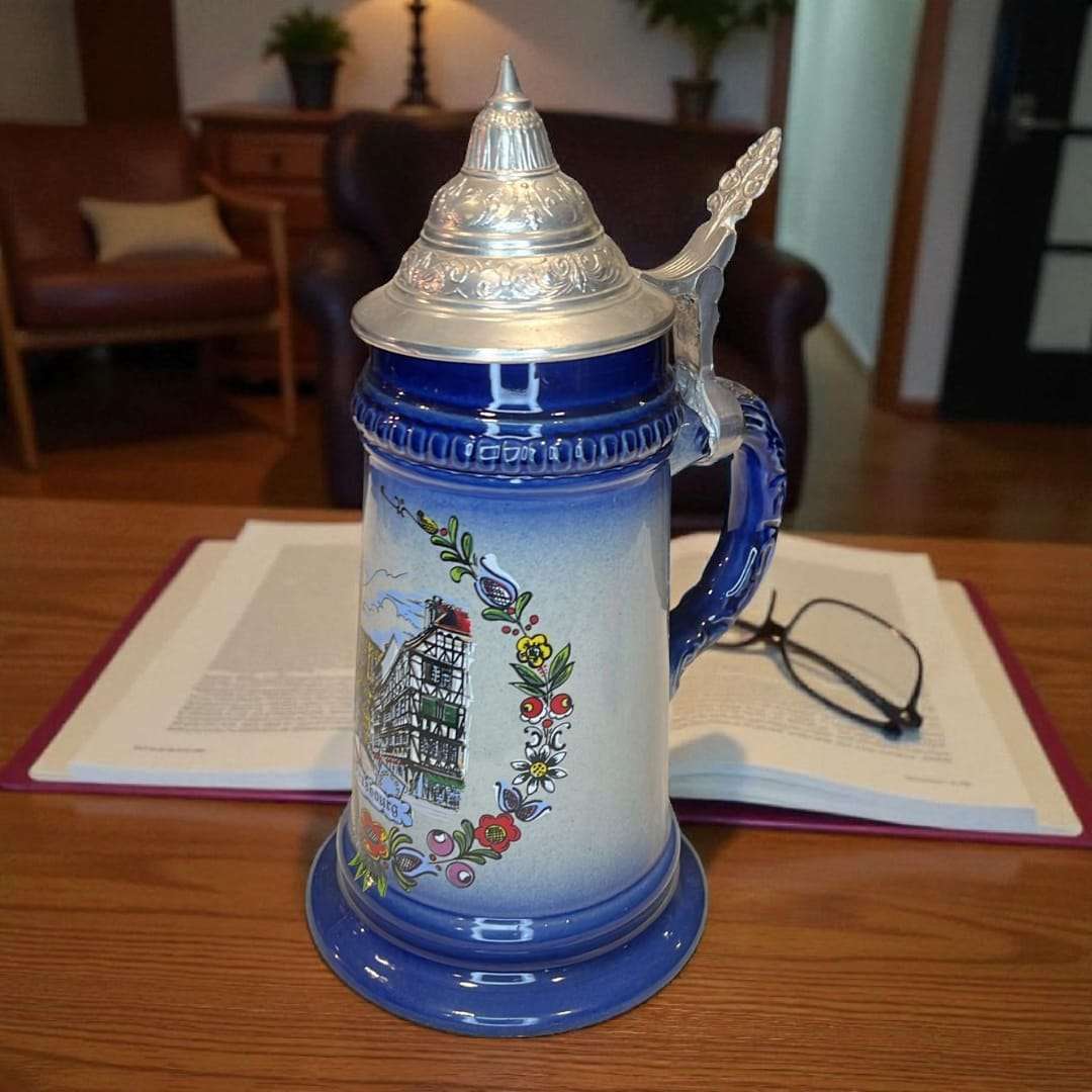 Jarra de cerveza alemana de porcelana con tapa metálica exclusiva, colocada sobre escritorio con libros, ideal para decoración clásica y temática europea.