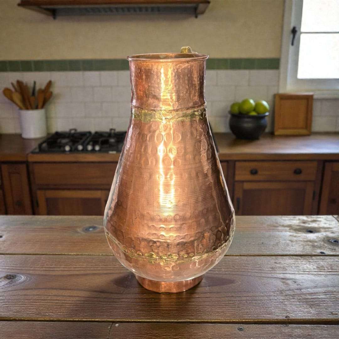 
La jarra de cobre con asa de metal destaca por su cuerpo abombado y cuello ligeramente estrechado, diseño pensado originalmente para conservar líquidos como agua