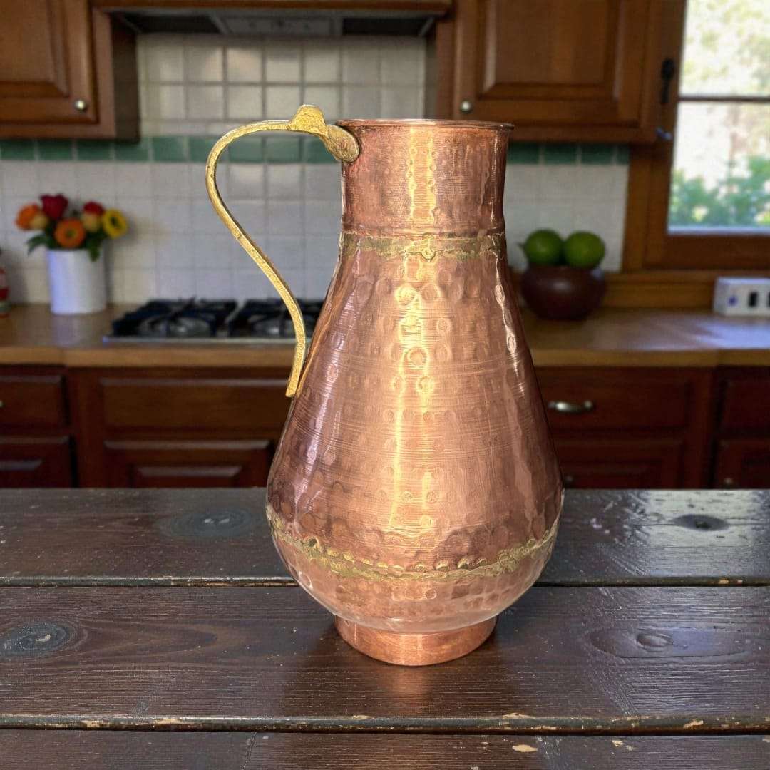 
La jarra de cobre con asa de metal destaca por su cuerpo abombado y cuello ligeramente estrechado, diseño pensado originalmente para conservar líquidos como agua
