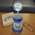 Jarra de cerveza alemana de porcelana con tapa metálica abierta, colocada sobre mesa con gafas y libro, perfecta para decoración de salas o bibliotecas.