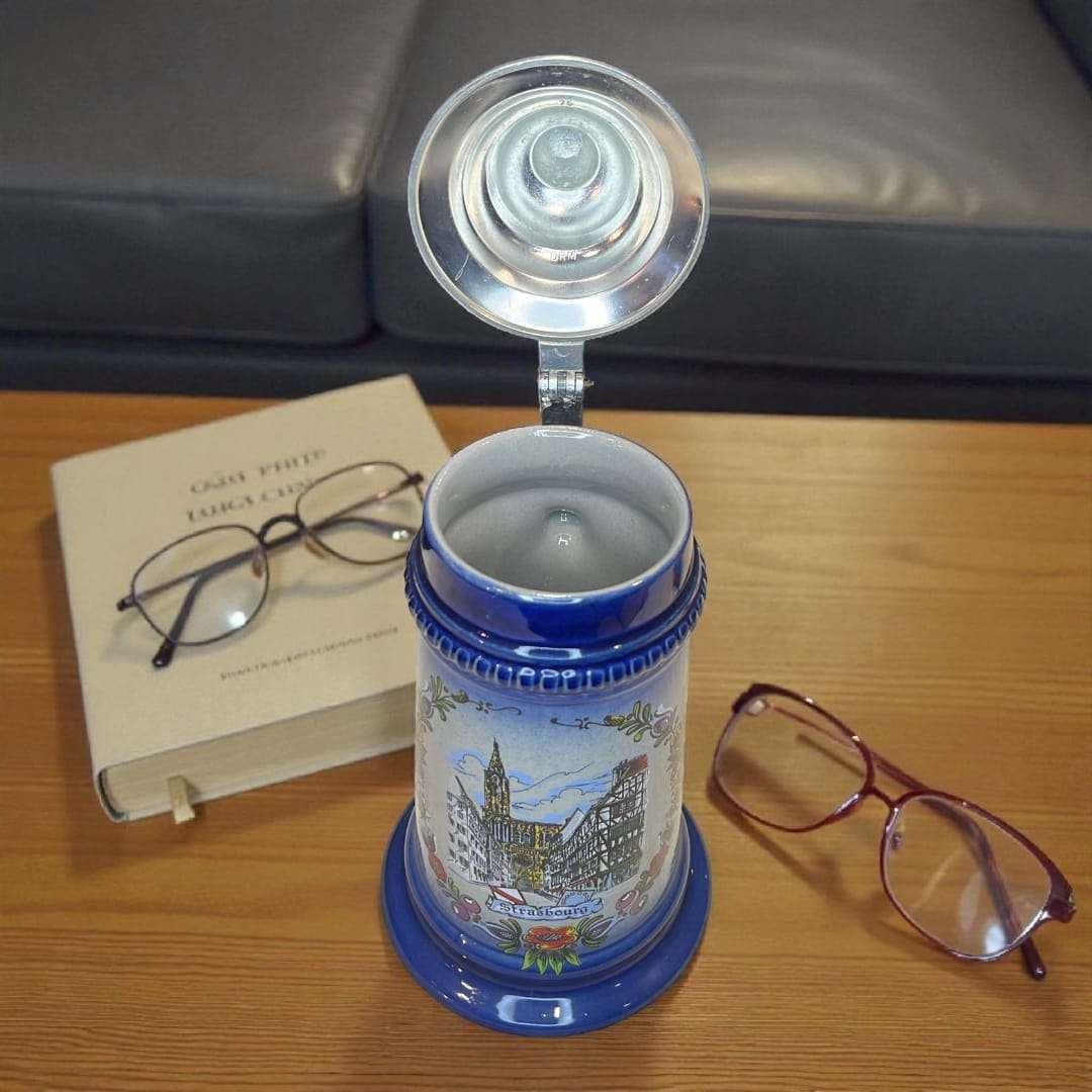 Jarra de cerveza alemana de porcelana con tapa metálica abierta, colocada sobre mesa con gafas y libro, perfecta para decoración de salas o bibliotecas.