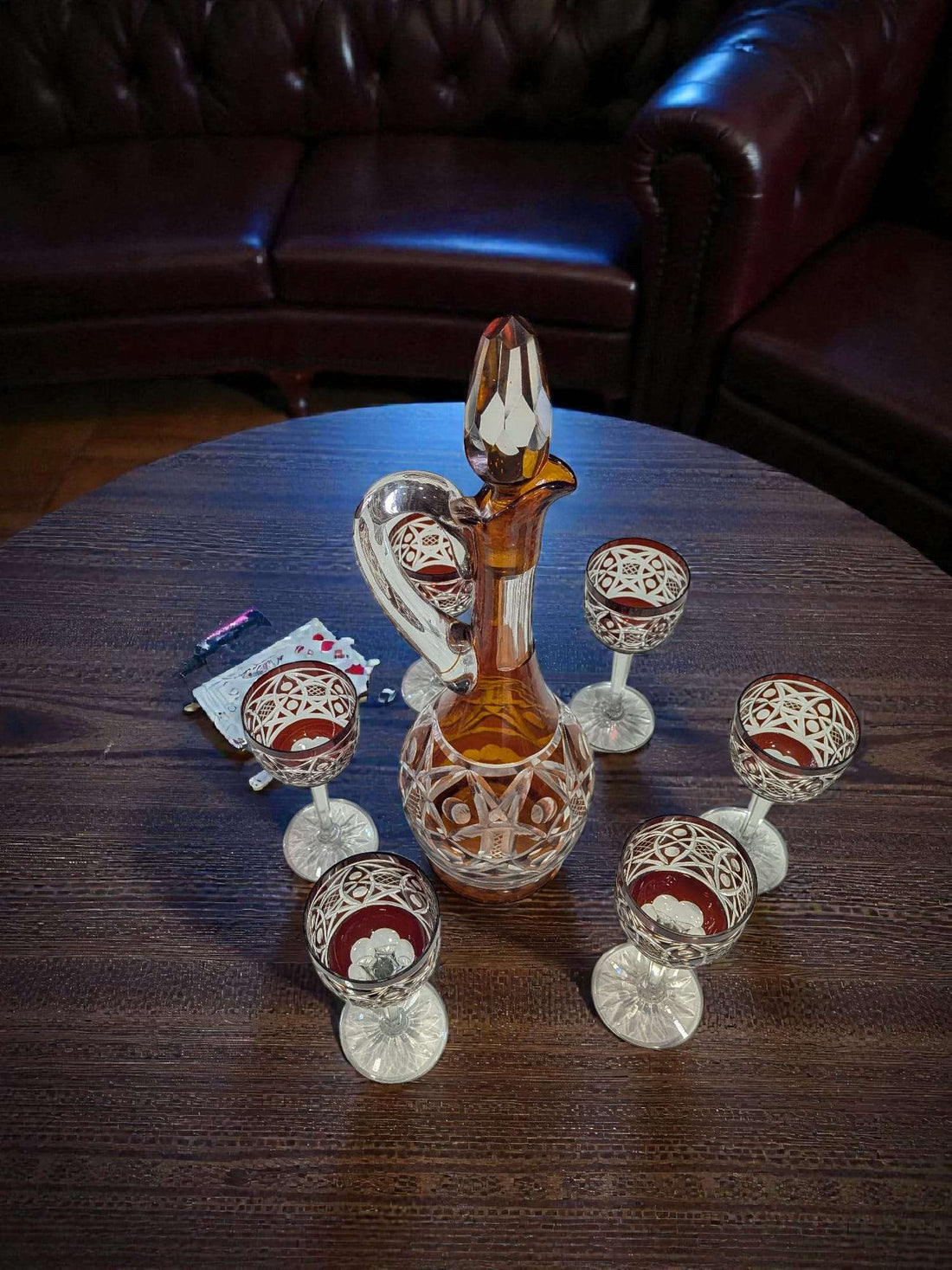 Juego de licorera con 6 copas de cristal tallado con color ámbar y rojo, sobre mesa de madera, ideal para decoración elegante de interiores.
