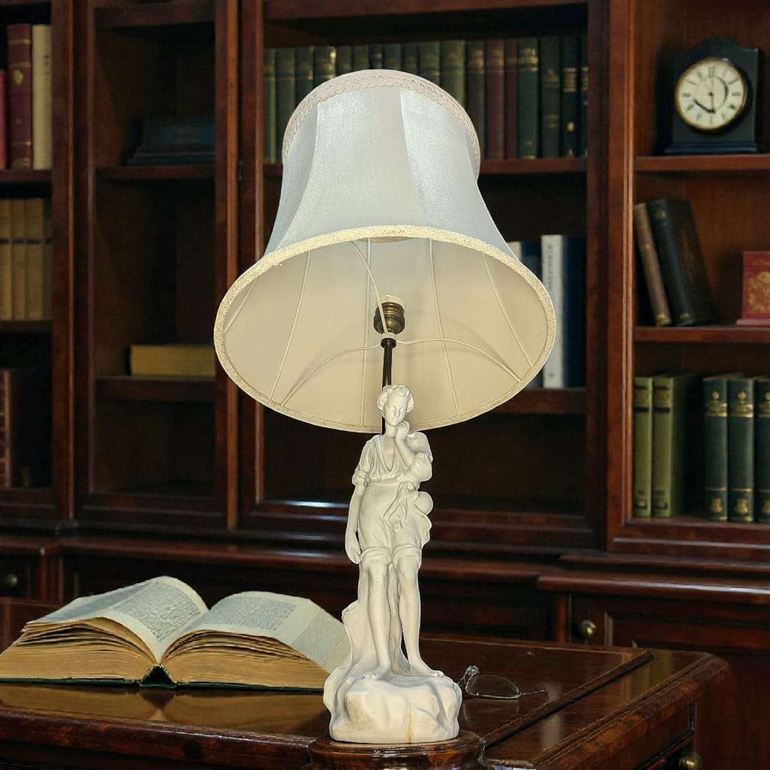 Lámpara de sobremesa con figura de porcelana biscuit de estilo clásico sobre peana, pantalla blanca y fondo de biblioteca elegante. Ideal para decoración vintage.