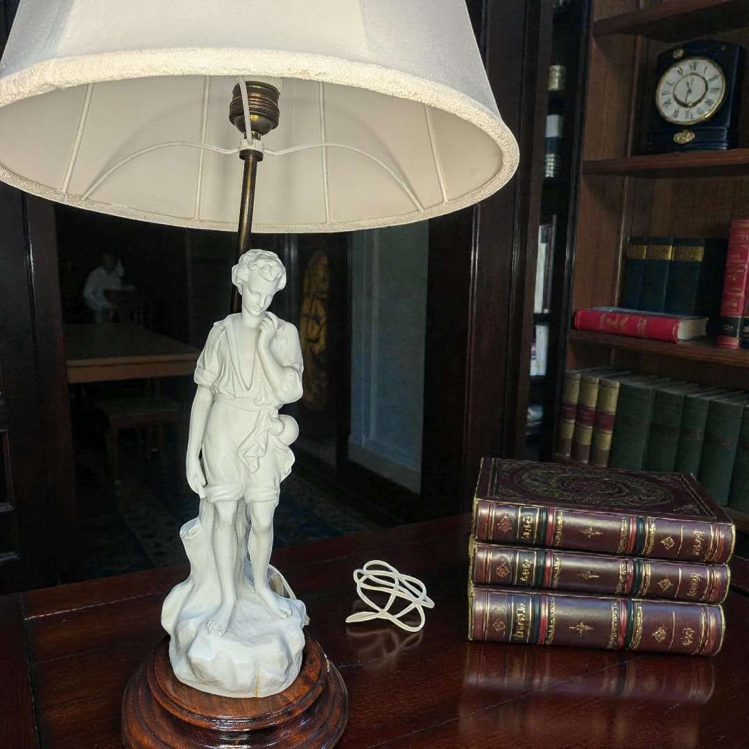 Lámpara de sobremesa con figura de porcelana biscuit sobre base de madera y pantalla blanca, ideal para decoración clásica en interiores elegantes.