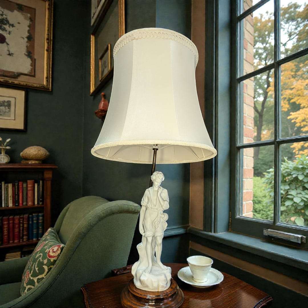 Lámpara de sobremesa con figura de porcelana biscuit sobre base de madera y pantalla blanca, ideal para decoración clásica en salones elegantes.