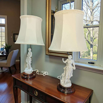 Lamparas de sobremesa con figuras de porcelana biscuit sobre peana de madera y pantalla blanca, ideales para decoración clásica en interiores elegantes.