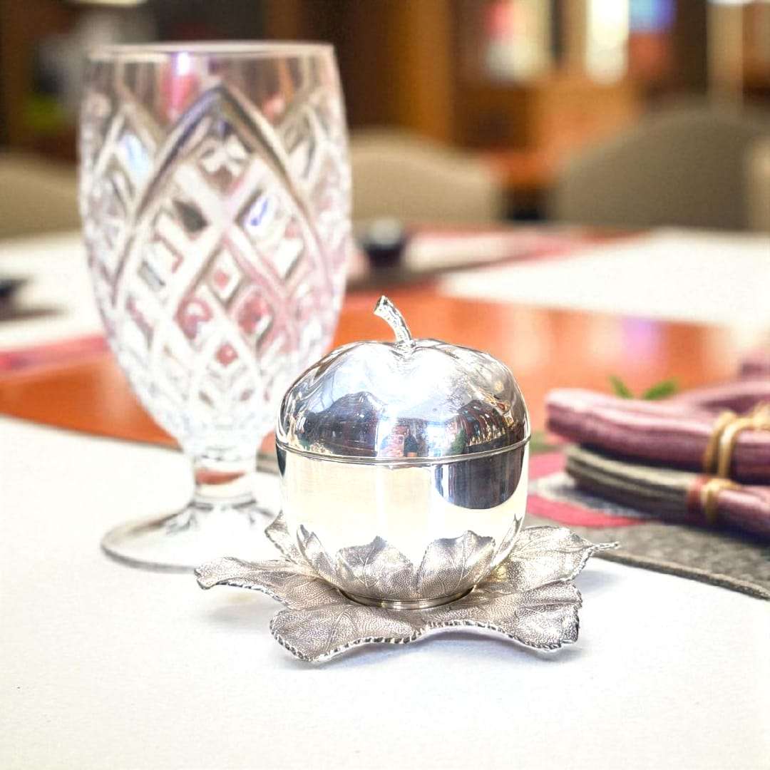 Azucarero de metal plateado con forma de manzana sobre base de hojas, junto a copa tallada, ideal para mesas refinadas y decoración exclusiva.