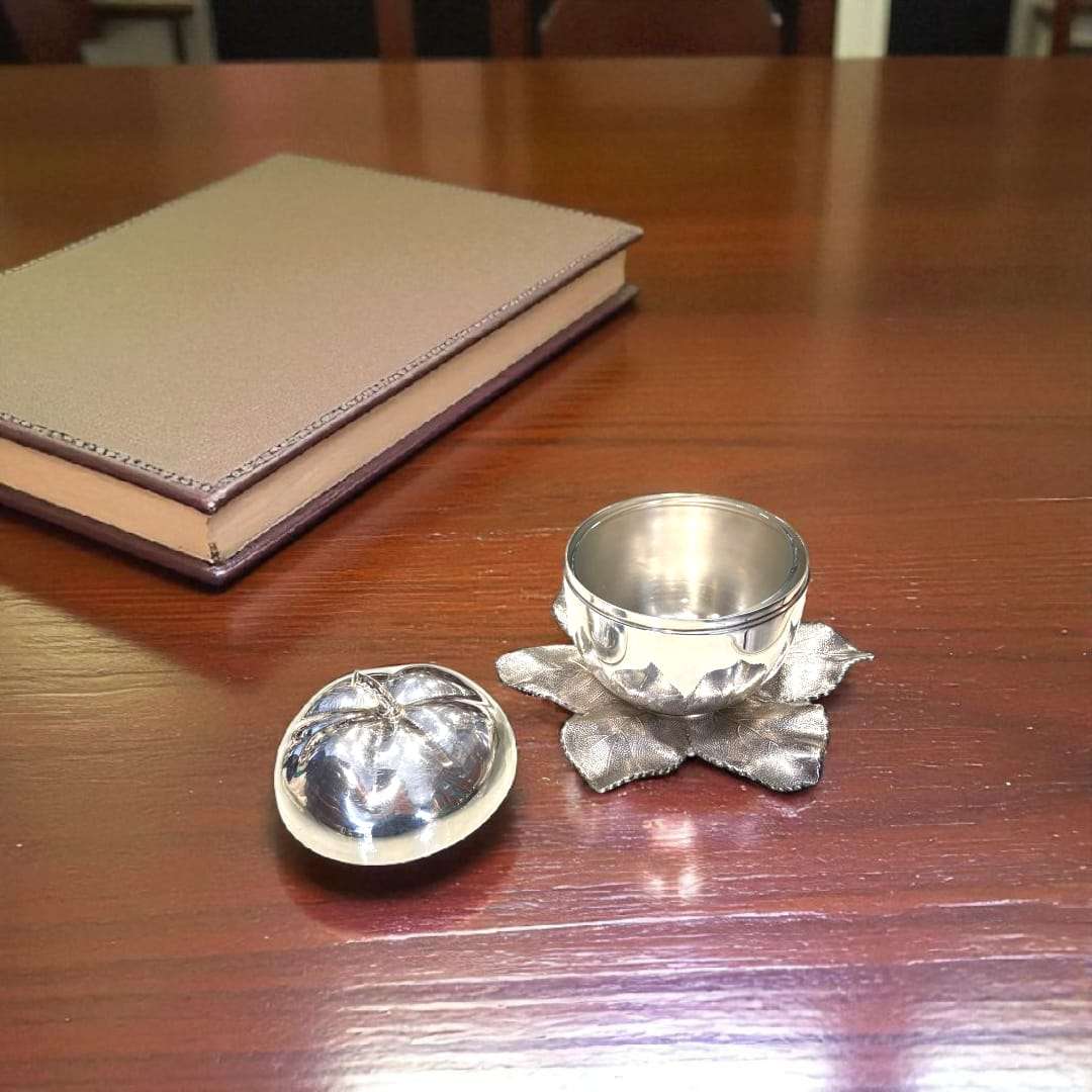 Azucarero de metal plateado con forma de manzana abierta, sobre base de hojas, en mesa de madera con libro decorativo, estilo sofisticado.