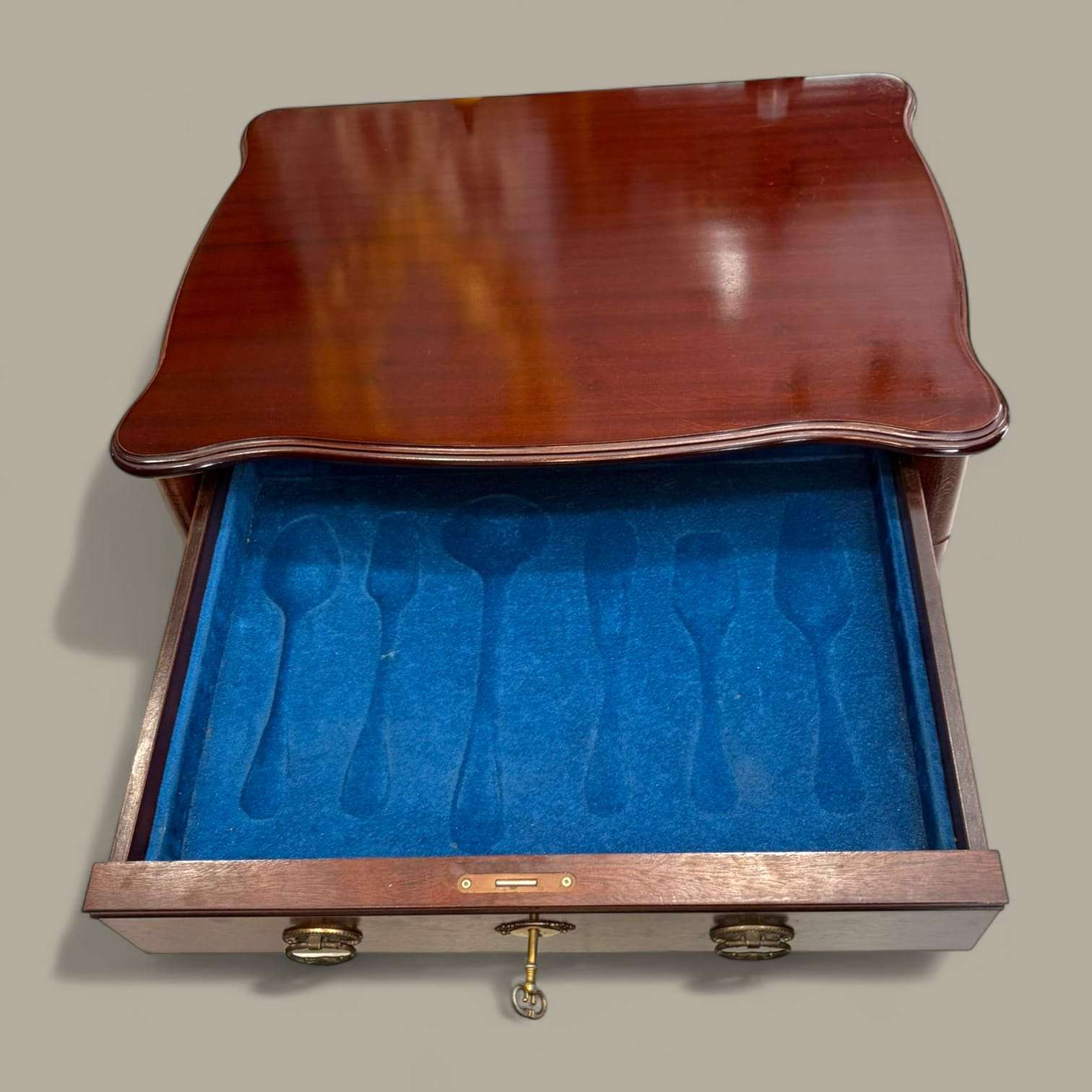 Mueble cubertero de madera con cajón abierto mostrando interior forrado en terciopelo azul con compartimentos para cubiertos. Incluye llave decorativa.