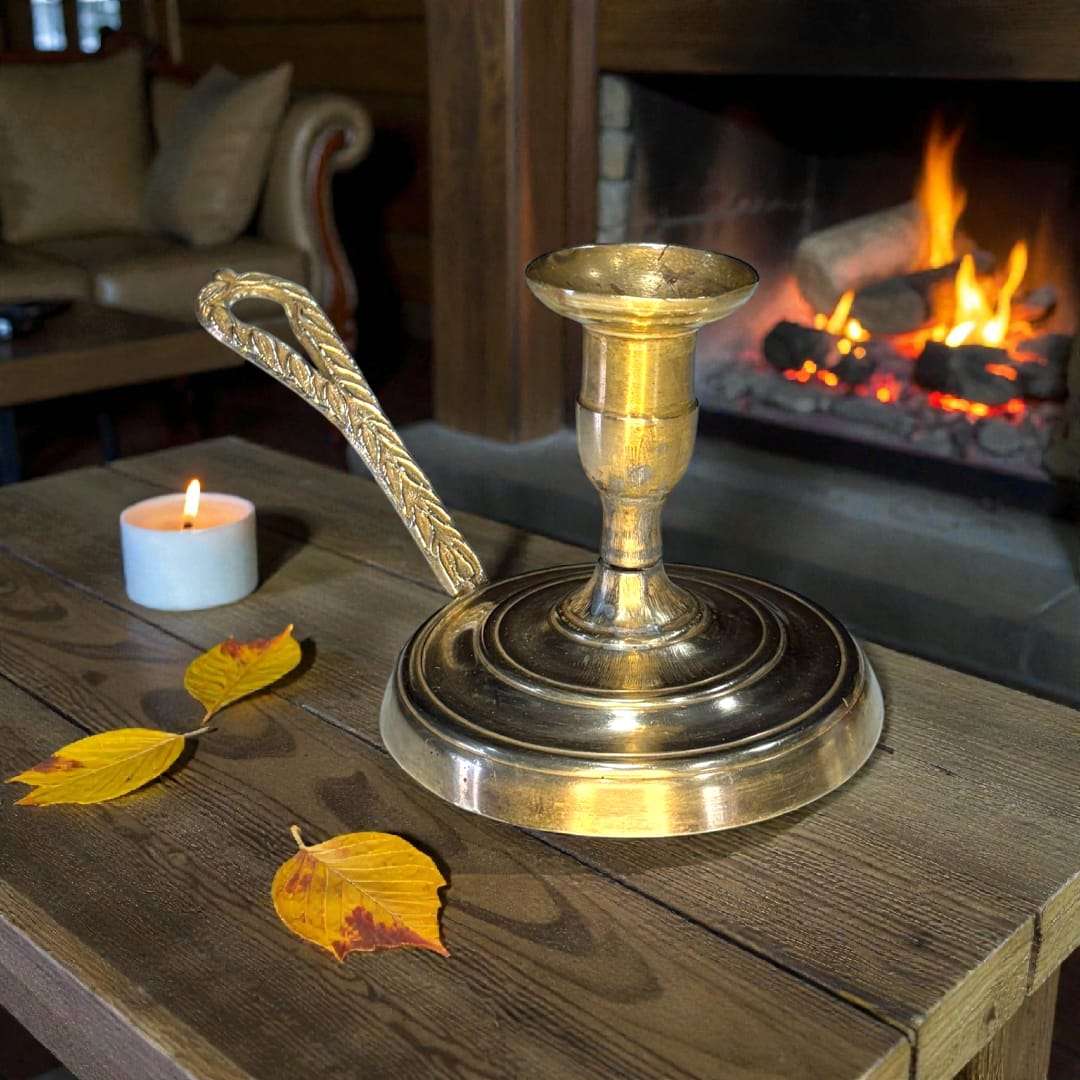 Palmatoria antigua de metal dorado con mango en relieve sobre mesa rústica frente a chimenea encendida, ideal para decoración clásica.