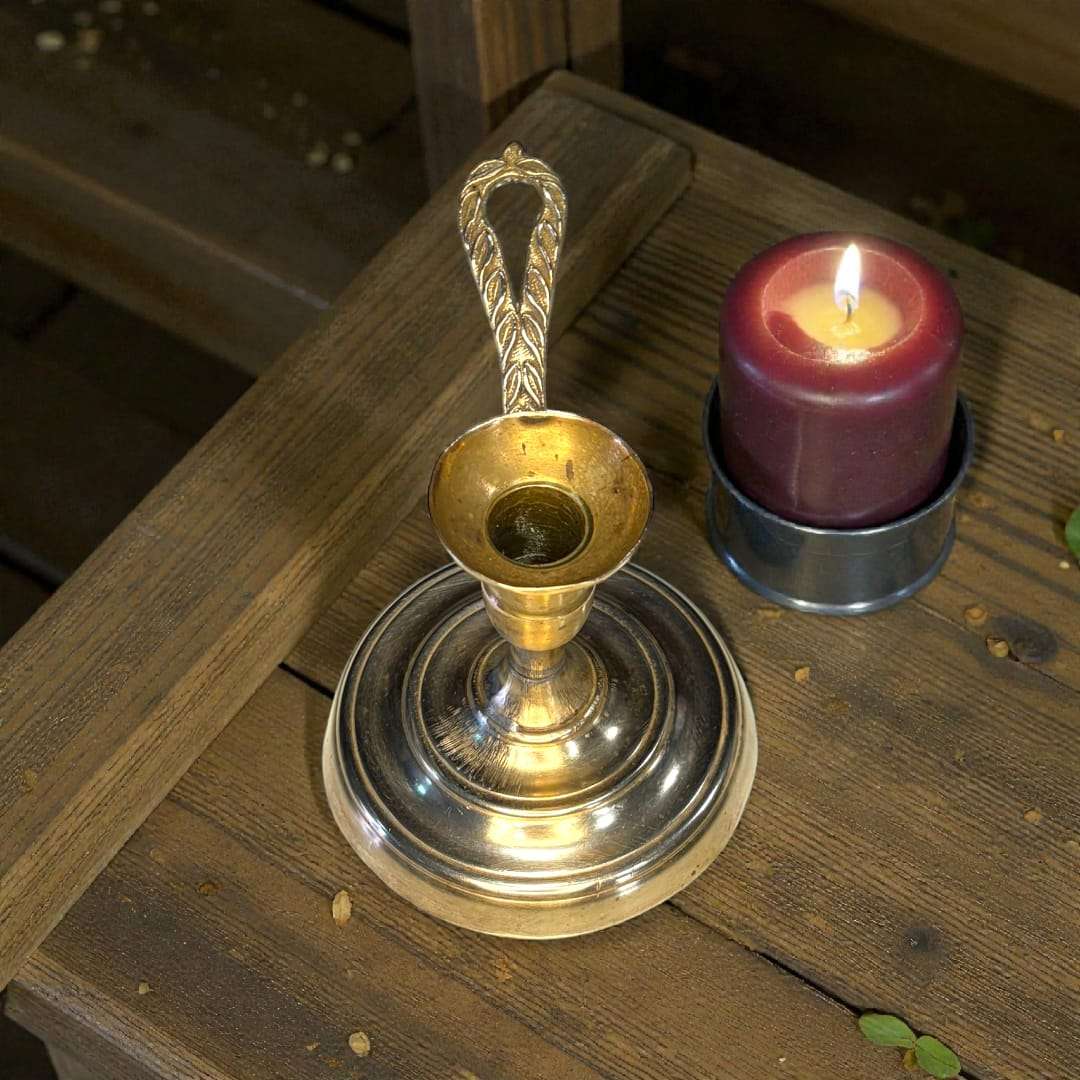 Palmatoria antigua de metal dorado con vela encendida al lado, destacando su mango en relieve y estilo decorativo clásico.