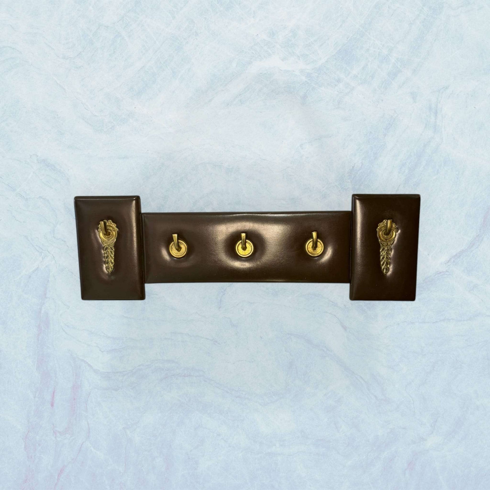 Perchero elegante de metal y cuero exclusivo y único con cuatro ganchos dorados sobre base marrón oscura. Ideal para decoración de interiores con estilo.
