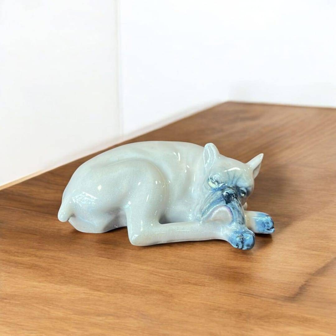 Perro de porcelana Leonardo PEYRO en posición de descanso, con detalles realistas y esmaltado azul en hocico y patas. Pieza exclusiva de decoración.