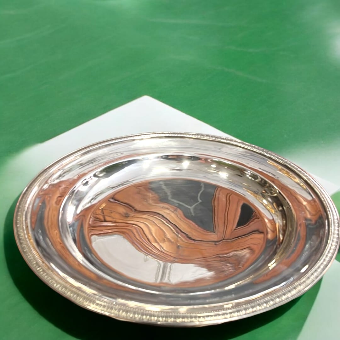 Bandeja redonda de metal plateado con diseño decorativo en el borde, sobre una superficie verde brillante.