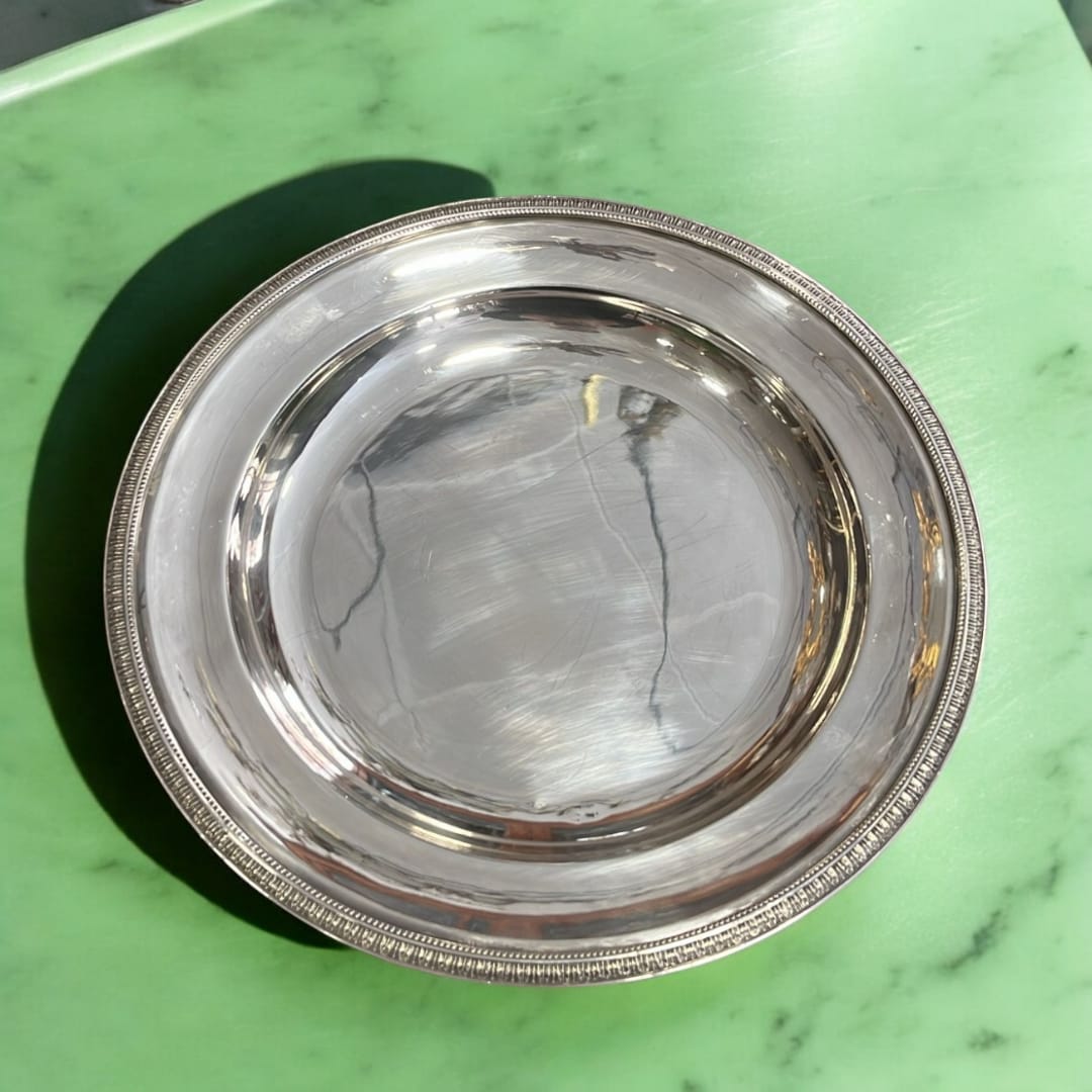 Bandeja redonda de metal plateado con diseño decorativo en el borde, sobre una superficie verde brillante.