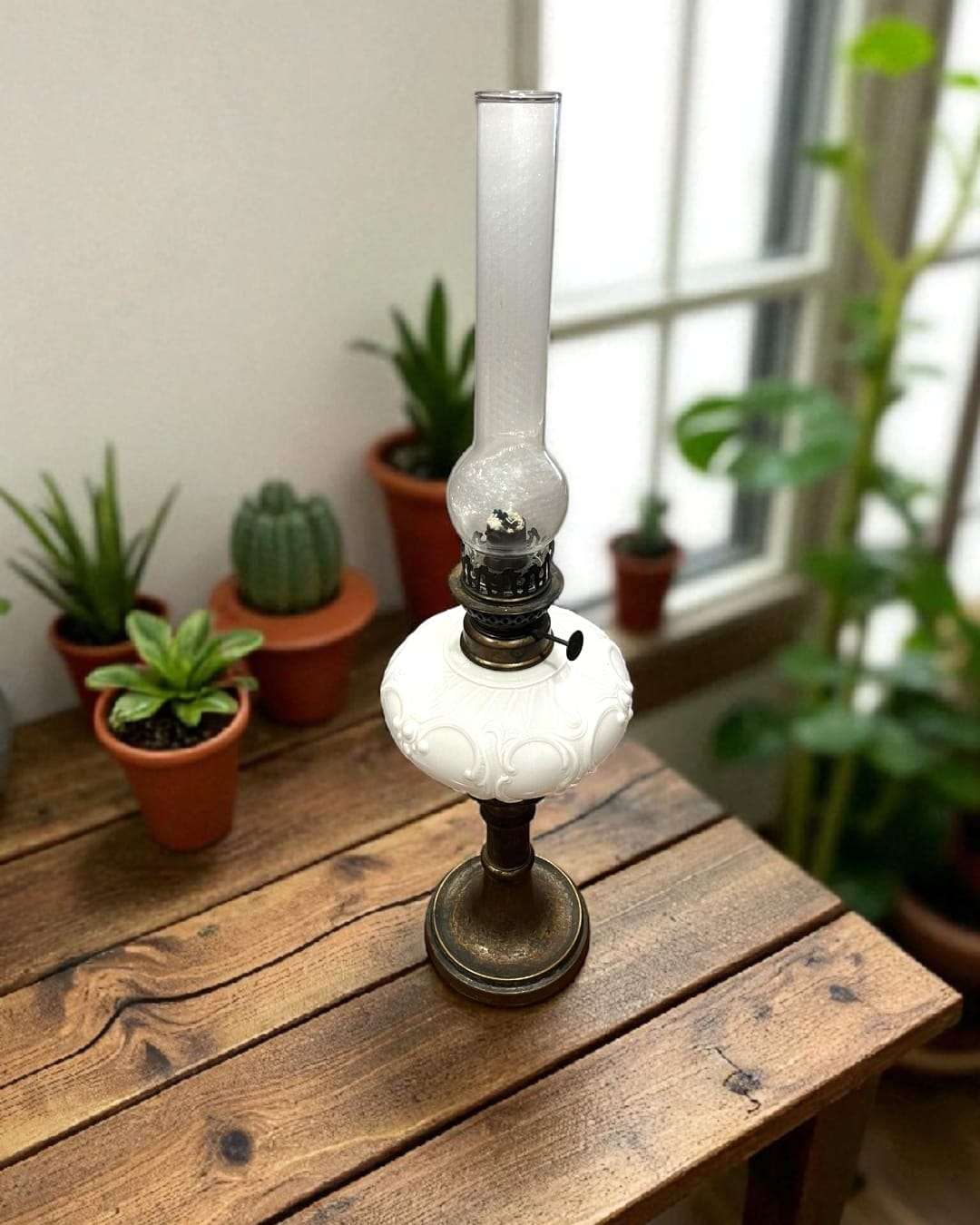 Quinqué vintage con base metálica y tulipa de cristal, colocado en mesa de madera con plantas, ideal para decoración de interiores.