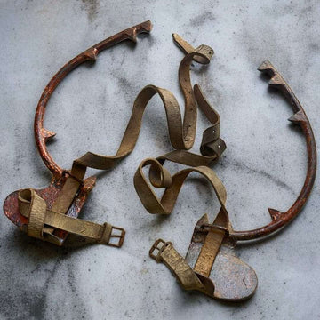 Par de trepadores antiguos de hierro forjado con correas de cuero en fondo gris concreto.